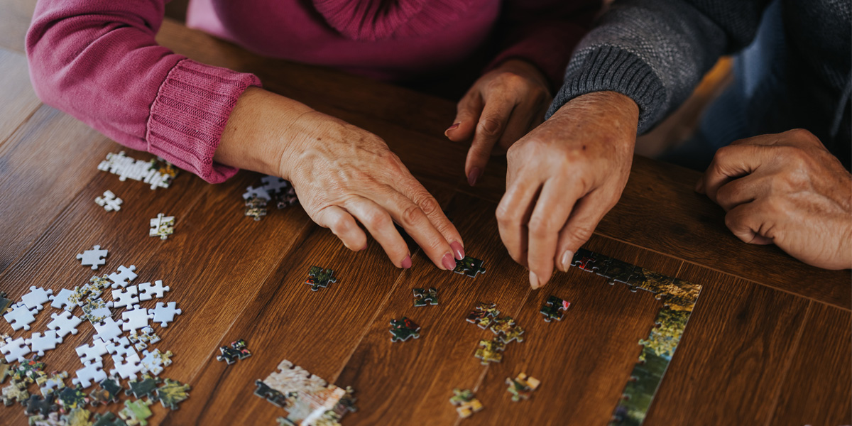 Close-up of two older adults working together on a jigsaw puzzle at a wooden table.
