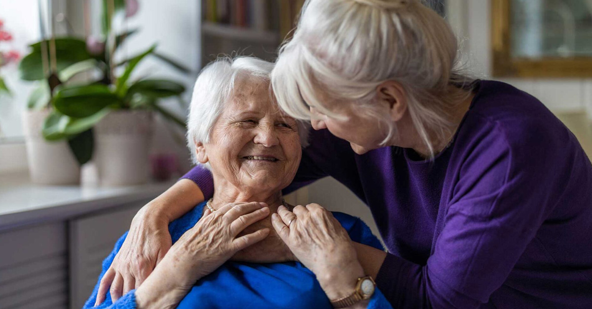 Older adult sitting indoors while a caregiver offers supportive touch, illustrating compassionate memory care and dementia support.