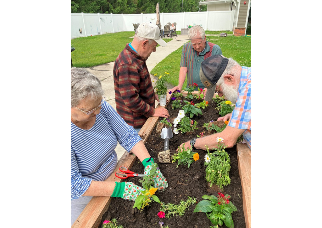 Residents at Meadowview of Johnston planting flowers together in a raised garden bed, using gardening tools in an outdoor courtyard with grass and patio seating nearby.