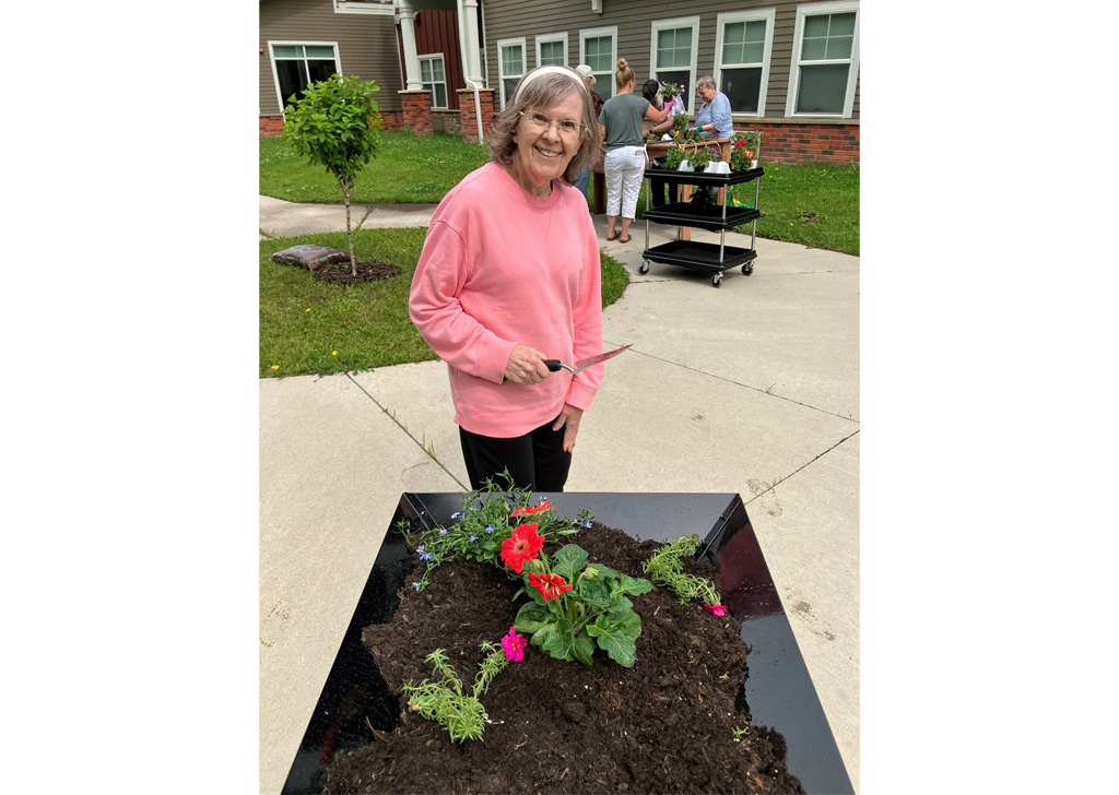Resident planting flowers in a raised garden bed at Meadowview of Johnston, holding a gardening tool while other residents and staff work together in the courtyard near the community building.
