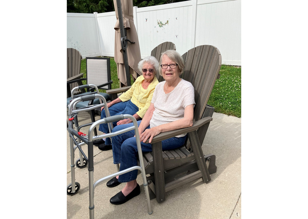 Residents seated outdoors on a patio at a senior living community, relaxing in Adirondack chairs near walkers, with a white fence and green lawn in the background.