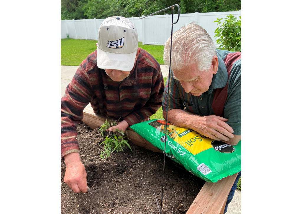 Residents gardening in a raised bed at a senior living community, planting in soil with a bag of garden soil nearby and a fenced outdoor patio in the background.