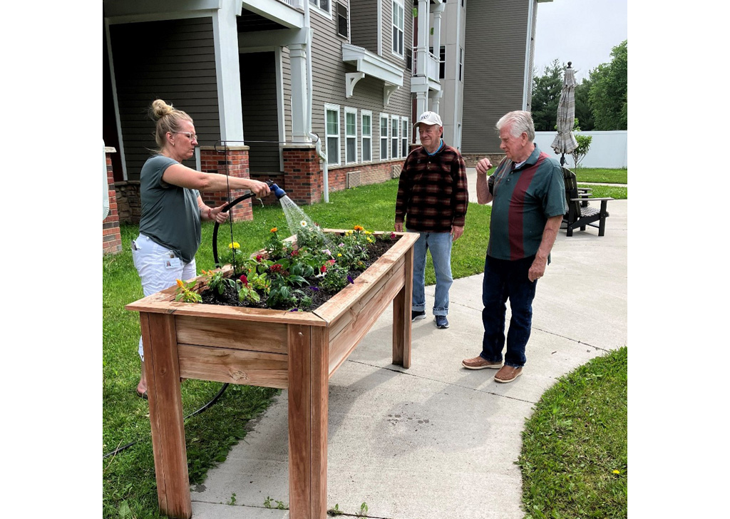 Staff member watering flowers in a raised garden bed at Meadowview of Johnston while residents observe and participate in an outdoor memory care gardening activity within the community courtyard.