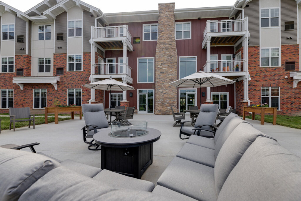 Outdoor fire pit patio at Meadowview of Johnston senior living community featuring comfortable seating, dining tables, and apartment balconies.
